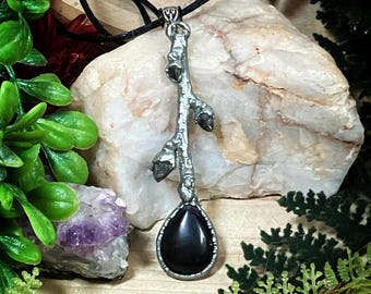May include: A silver-toned pendant necklace featuring a branch-like design with three small, dark-colored stones and a large, teardrop-shaped black stone. The pendant hangs from a black cord, set against a natural background of rocks and greenery.