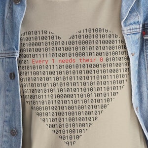 Może przedstawiać: Beżowa koszulka z sercowatym wzorem wykonanym z kodu binarnego. Tekst "Every 1 needs their 0" jest nadrukowany na czerwono wewnątrz serca.