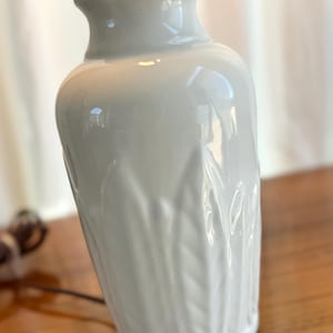 Puede incluir: Una lámpara de mesa de cerámica blanca con un diseño de hojas texturizadas. La lámpara tiene una base redondeada y una parte superior acampanada, con una tapa de metal plateado. La lámpara está sobre una superficie de madera.