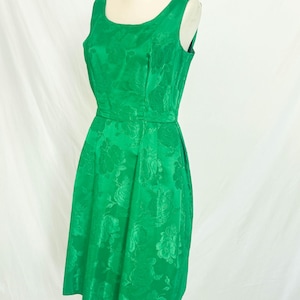 May include: A sleeveless, knee-length green dress with a floral pattern. The dress features a scoop neckline, fitted bodice, and a flared skirt. The fabric has a subtle sheen, and the dress is displayed on a mannequin.