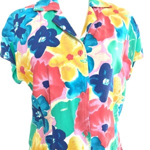 May include: A short-sleeved button-down shirt featuring a vibrant, large-scale floral print in shades of bright pink, yellow, teal, and dark blue. The garment has a classic collar, a straight hem, and three visible red buttons down the front centre.