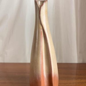 May include: A beige and peach ceramic vase with a distinctive, twisted form. The vase features a narrow neck and a broader base, with a colour gradient from beige to peach. The vase is a decorative item, suitable for display.