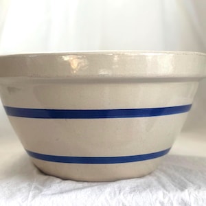 Puede incluir: Un cuenco mezclador de gres beige con dos rayas azules horizontales. El cuenco tiene forma redondeada y un borde ancho. Este utensilio de cocina vintage es adecuado para mezclar ingredientes.