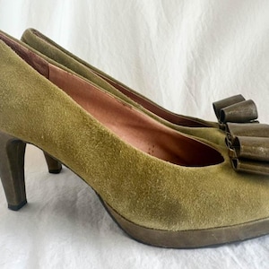 May include: Olive green suede high heels with a platform sole and a dark brown bow detail on the toe. The heels have a rounded toe and a dark brown heel. The shoes are a size 8.