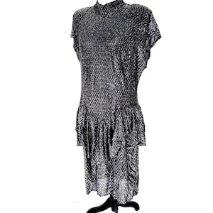 May include: A metallic silver and black textured dress featuring a high neckline, short sleeves, and a dropped waistline with a ruffled skirt detail. The garment has a shimmering, open-weave fabric construction that creates a distinct grid-like pattern.