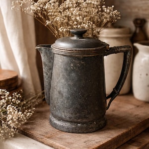 May include: An antique, dark gray metal coffee pot with a lid and handle, set on a wooden surface. The pot is surrounded by dried white flowers, creating a rustic, vintage aesthetic. Other ceramic items are in the background.