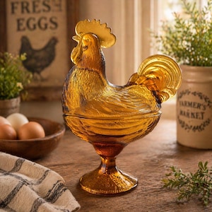 May include: Amber-colored glass rooster-shaped decorative dish with a textured surface. The dish has a pedestal base and a lid. A wooden bowl of eggs and a sign that says "FRESH EGGS" are in the background.