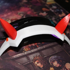 Puede incluir: Un accesorio para ratón de ordenador impreso en 3D de color blanco y rojo con dos cuernos. El accesorio está diseñado para colocarse sobre un ratón de ordenador.