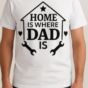 Puede incluir: Camiseta blanca con un diseño gráfico negro de una casa con una estrella en la parte superior. El texto dice "Home is where Dad is" con dos símbolos de llave inglesa a cada lado del texto.