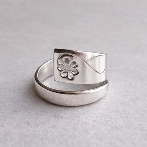Puede incluir: Un anillo de plata con un diseño floral. El anillo presenta una parte superior rectangular con una flor recortada y un diseño curvo. La banda es de plata pulida simple. El anillo se muestra sobre una superficie gris claro.
