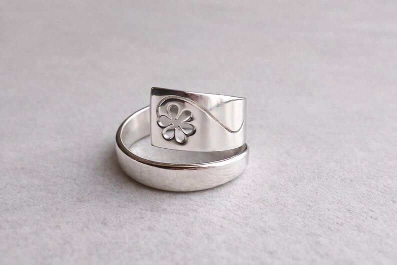 Op de afbeelding: Een zilveren ring met een bloemmotief. De ring heeft een rechthoekige bovenkant met een uitgesneden bloem en een gebogen ontwerp. De band is eenvoudig, gepolijst zilver. De ring wordt gepresenteerd op een lichtgrijze ondergrond.