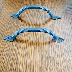 Peut inclure: Deux poignées en métal gris foncé torsadé avec des extrémités en forme de feuille. Les poignées sont arquées et semblent être en fer forgé. Elles sont posées sur un fond à grain de bois.