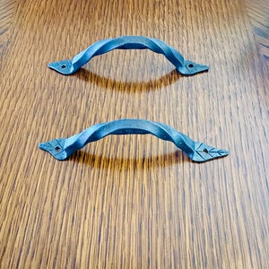 Peut inclure: Deux poignées de meuble en métal torsadé gris foncé. Chaque poignée a un design incurvé et arqué avec des extrémités décoratives en forme de feuille. Les poignées sont posées sur un fond de grain de bois.