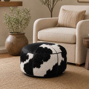 May include: A round, low-profile ottoman featuring a black and white cowhide pattern with visible hair texture. The piece is constructed with distinct vertical seams and a smooth black piping border along the top and bottom edges of the circular frame.