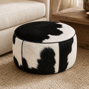 May include: A round ottoman featuring a black and white cowhide pattern with a textured hair-on-hide finish. The cylindrical piece is constructed with distinct vertical seams and dark piping along the top edge, creating a structured profile.