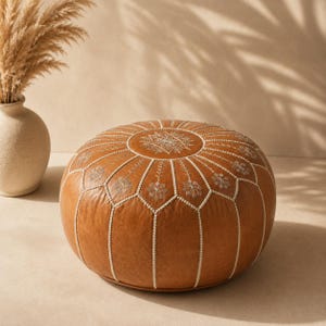 May include: A round, tan leather pouf featuring white stitched seams and intricate white embroidered floral patterns on the top and sides. The pouf has a structured, low-profile design with a smooth surface texture and visible decorative stitching.