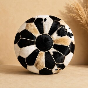May include: Circular ottoman with a cowhide pattern in black, white, and tan. The design features a central black circle surrounded by geometric shapes. The edges are detailed with small, white beads. The ottoman is approximately 76 cm in diameter.