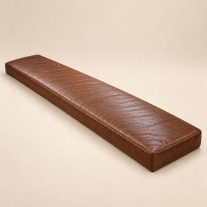 May include: A long, rectangular bench cushion upholstered in a smooth, medium-brown material with a subtle grain texture. The cushion features clean, structured edges and a flat, uniform surface, presenting a simple, elongated form.