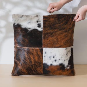 Peut inclure: Coussin carré décoratif avec un motif patchwork. Le coussin présente quatre sections en peau de vache dans les tons marron, noir et blanc. Le coussin est tenu par deux mains.