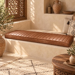May include: A long, rectangular bench cushion upholstered in smooth, tan-colored faux leather. The cushion features a series of vertical channel-stitched lines running across its entire length, creating a uniform, ribbed texture on the surface.
