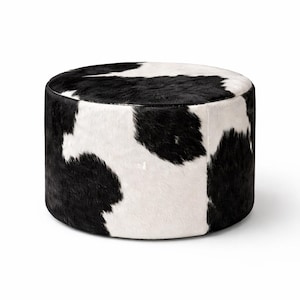 May include: A round ottoman with a cowhide pattern. The ottoman features a black and white spotted design, with the black spots irregularly shaped against a white background. The sides are a solid black color.