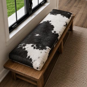 May include: A rectangular wooden bench featuring a long, cushioned seat upholstered in a black and white cowhide pattern. The bench has a sturdy, natural-toned wooden frame with four legs and a flat, horizontal surface supporting the cushion.