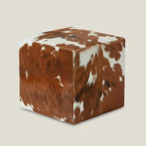 Könnte beinhalten: Ein würfelförmiger Hocker mit Kuhfellmuster. Der Hocker hat ein braun-weißes Fleckenmuster, wobei die braunen Bereiche die Oberfläche dominieren. Der Würfel ist ein neutrales Akzentstück für jeden Raum.