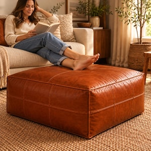 May include: A large, square, cognac-colored leather ottoman featuring a smooth, matte finish and visible stitched grid detailing across its top and sides. The ottoman has a low-profile, boxy shape and rests directly on a textured, woven floor surface.