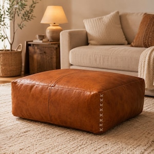 Peut inclure: Un pouf de sol carré à profil bas recouvert de cuir couleur cognac. La pièce présente une surface supérieure lisse et sans couture avec une couture centrale et des détails de points en croix clairs visibles sur les bords latéraux verticaux.