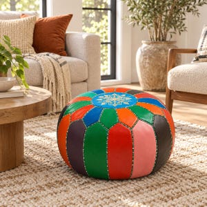 May include: A round, multi-colored leather pouf featuring vertical panels in shades of red, orange, green, blue, pink, and dark brown. The top center displays a circular blue panel with a white, intricate, star-like geometric pattern.