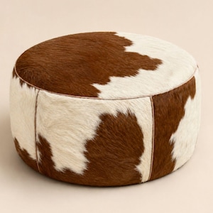 May include: A round ottoman upholstered in genuine cowhide featuring a natural brown and white spotted pattern. The cylindrical piece is constructed with visible vertical seams and a smooth, low-profile design, showcasing the textured hair.
