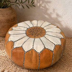 Peut inclure: Pouf rond en cuir, de couleur beige et crème. Le dessus présente un motif géométrique marron et crème, avec des coutures noires. Un élément décoratif pour un salon ou une chambre. Environ 60 cm de diamètre.
