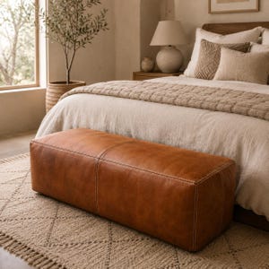 Op de afbeelding: Een rechthoekige, cognac-kleurige lederen bank met zichtbare witte stiksels langs de naden. De bank heeft een gladde, matte afwerking en een gestructureerd, laag ontwerp, horizontaal geplaatst voor een bed in een slaapkamer.