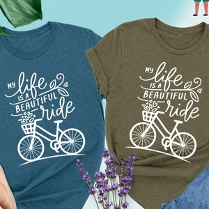 May include: Two t-shirts with the text "My life is a beautiful ride" and a bicycle graphic. The shirt on the left is teal and the shirt on the right is olive.