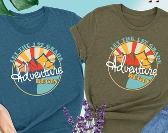 Puede incluir: Dos camisetas con un diseño gráfico retro. El diseño presenta una cordillera con un sol y el texto "Let the 1st Grade Adventure Begin". Una camiseta es azul teal y la otra es verde oliva.