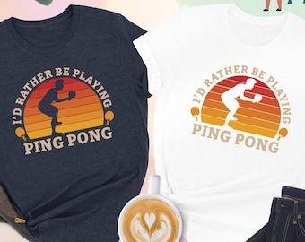 May include: Two t-shirts with a retro sunset graphic and a silhouette of a person playing ping pong. The text on the shirts reads "I'd rather be playing ping pong".