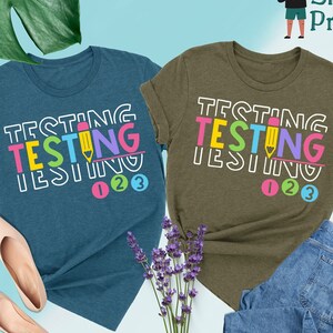 May include: Two t-shirts with the text "TESTING" in colorful letters and a pencil graphic. The shirts are in heather deep teal and heather olive colors. The text "123" is below the word "TESTING" on each shirt.