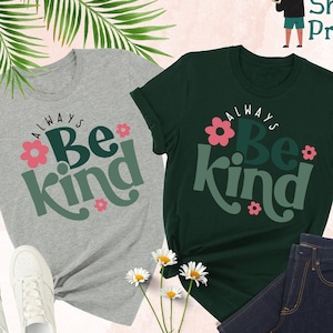 May include: Two t-shirts with the text "Always Be Kind" in green and pink lettering with pink flower accents. The shirts are on a white background with green leaves, white flowers, and a pair of white sneakers.