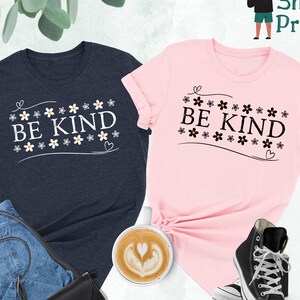 K&ouml;nnte beinhalten: Zwei T-Shirts mit dem Text "BE KIND" umgeben von kleinen Blumengrafiken. Die Shirts sind in Heather Navy und Pink.