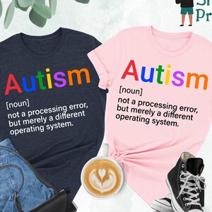 May include: Two t-shirts with the word "Autism" printed in rainbow colors. The text below the word defines autism as "not a processing error, but merely a different operating system". One shirt is heather navy and the other is pink.