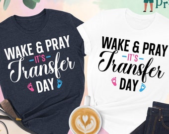 May include: Two t-shirts with the text "Wake & Pray - It's Transfer Day" printed on them. One shirt is heather navy and the other is white. Both shirts feature a pink and blue footprint graphic.