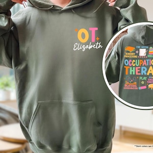May include: A military green hooded sweatshirt with a pocket on the front. The front of the sweatshirt has the text "OT Elizabeth" in pink, orange, and yellow. The back of the sweatshirt has the text "Occupational Therapy" in colorful text with icons.