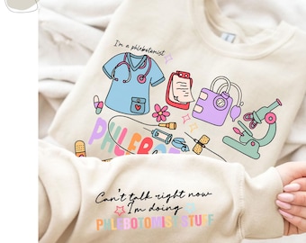 May include: A beige sweatshirt with a colourful graphic design featuring a cartoon phlebotomist and medical equipment. The text "I'm a phlebotomist" and "Can't talk right now I'm doing PHLEBOTOMIST STUFF" is printed on the sweatshirt.