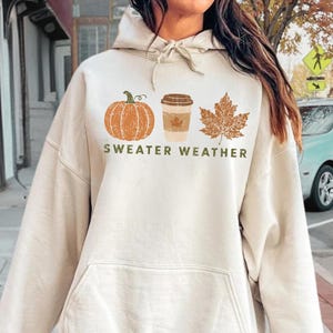 May include: A sand-colored hooded sweatshirt featuring a graphic print of an orange pumpkin, a coffee cup, and a brown maple leaf, with the text "SWEATER WEATHER" printed in dark green letters below the icons. The hoodie has a front kangaroo pocket.