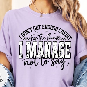 May include: An orchid-colored short-sleeve t-shirt featuring black text that reads I DON'T GET ENOUGH CREDIT For The Things I MANAGE not to say, with small star accents. The shirt is worn with a denim jacket and distressed denim shorts.
