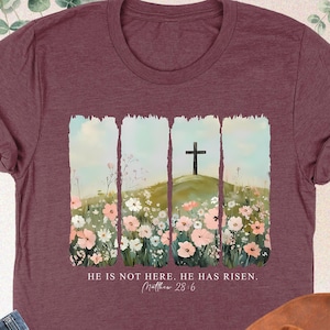 May include: Heather maroon t-shirt with a floral design and a cross on a hill. The text "He is not here. He has risen. Matthew 28:6" is printed on the shirt.
