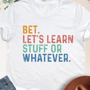 May include: White t-shirt with the text "BET. LET'S LEARN STUFF OR WHATEVER" in orange, red, green, and blue. The image also shows blue jeans and a brown fedora hat.