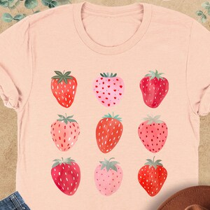 May include: A heather peach t-shirt with a graphic of nine red and pink strawberries. The strawberries are arranged in a 3x3 grid.