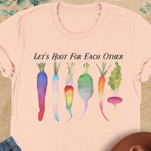 May include: A light pink t-shirt with the text "Let's Root For Each Other" and a watercolor illustration of colorful carrots, radishes, and turnips.