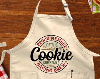 May include: A white apron with a pocket and a red, green, and black circle design that says "Proud Member of the Cookie Christmas Baking Crew".